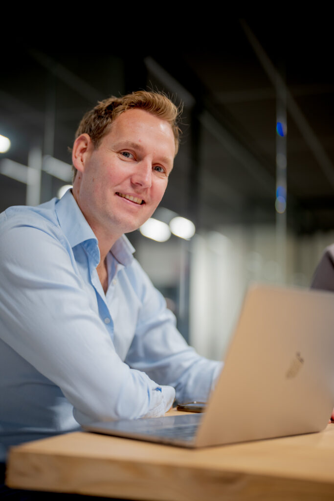 Een man in een lichtblauwe blouse zit aan een houten tafel met een laptop voor zich. Achter hem zijn glazen wanden en verlich