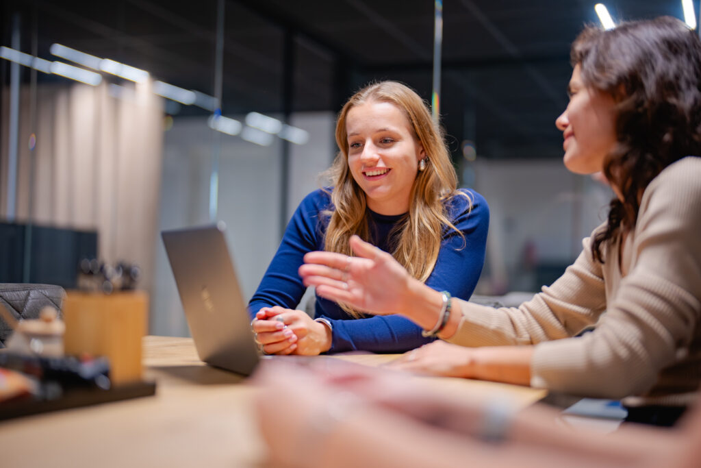 Twee vrouwen zitten aan een houten tafel met een laptop. Ze zijn betrokken in een gesprek. Op tafel staan pennen en notitiebl