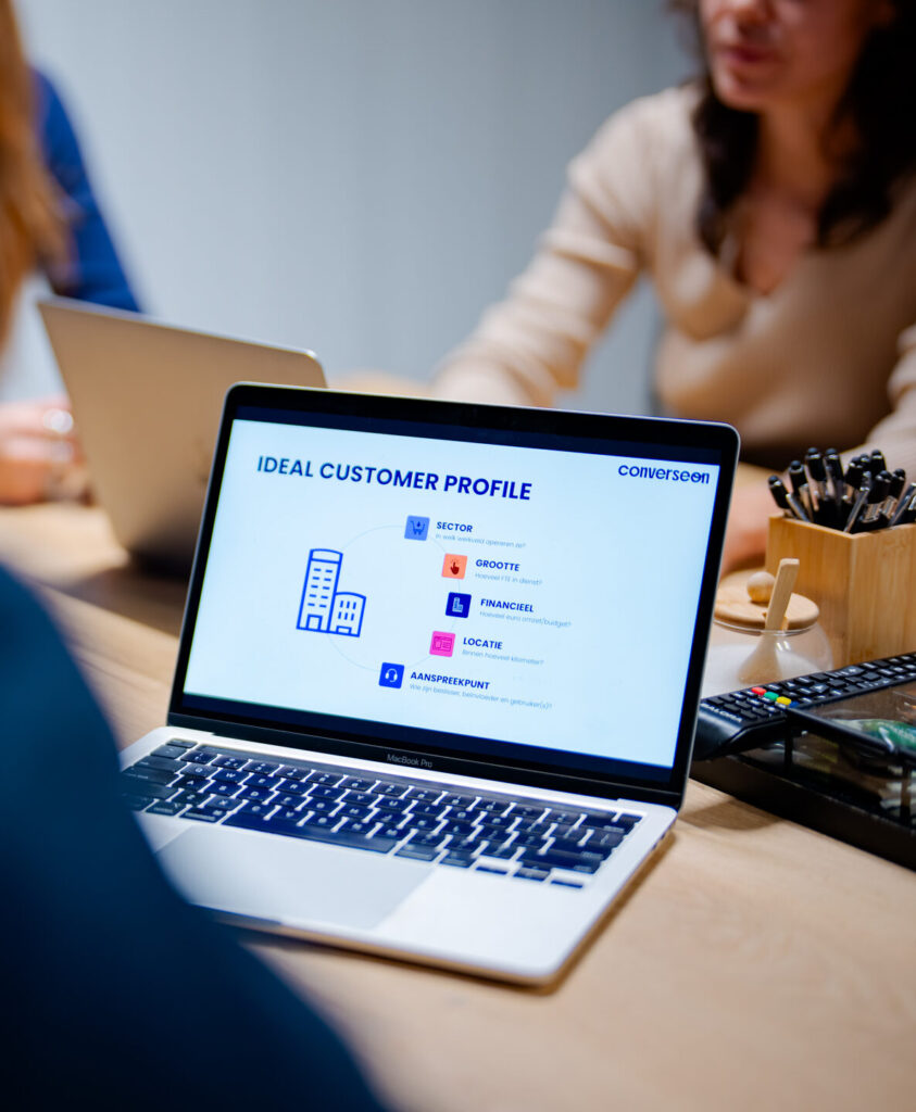 Een laptop staat open op een tafel met een presentatie over een ideaal klantenprofiel. Op de achtergrond zijn twee mensen te