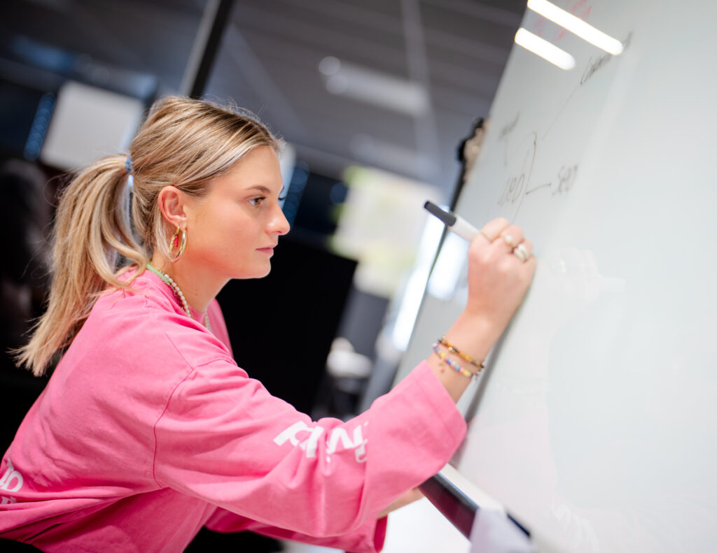 Een vrouw in een roze shirt schrijft met een stift op een whiteboard. De omgeving lijkt een moderne kantoorruimte met donkere
