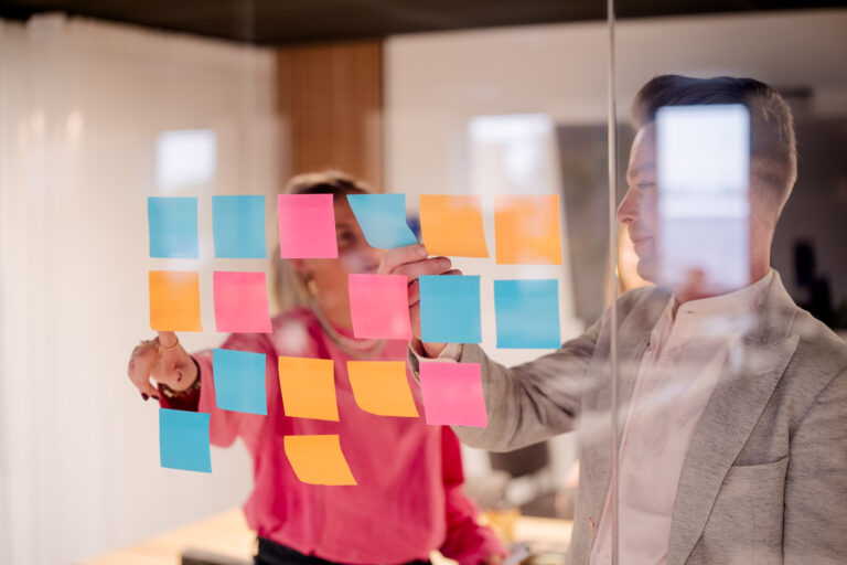 Twee mensen plakken gekleurde post-its op een glazen muur in een moderne kantooromgeving. De post-its zijn in verschillende k