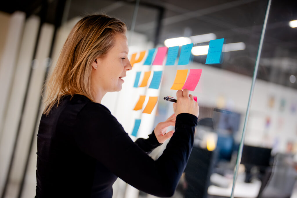 Een vrouw schrijft met een pen op een plastic oppervlak. Achter haar zijn kleurige post-its aangebracht. De omgeving lijkt ee