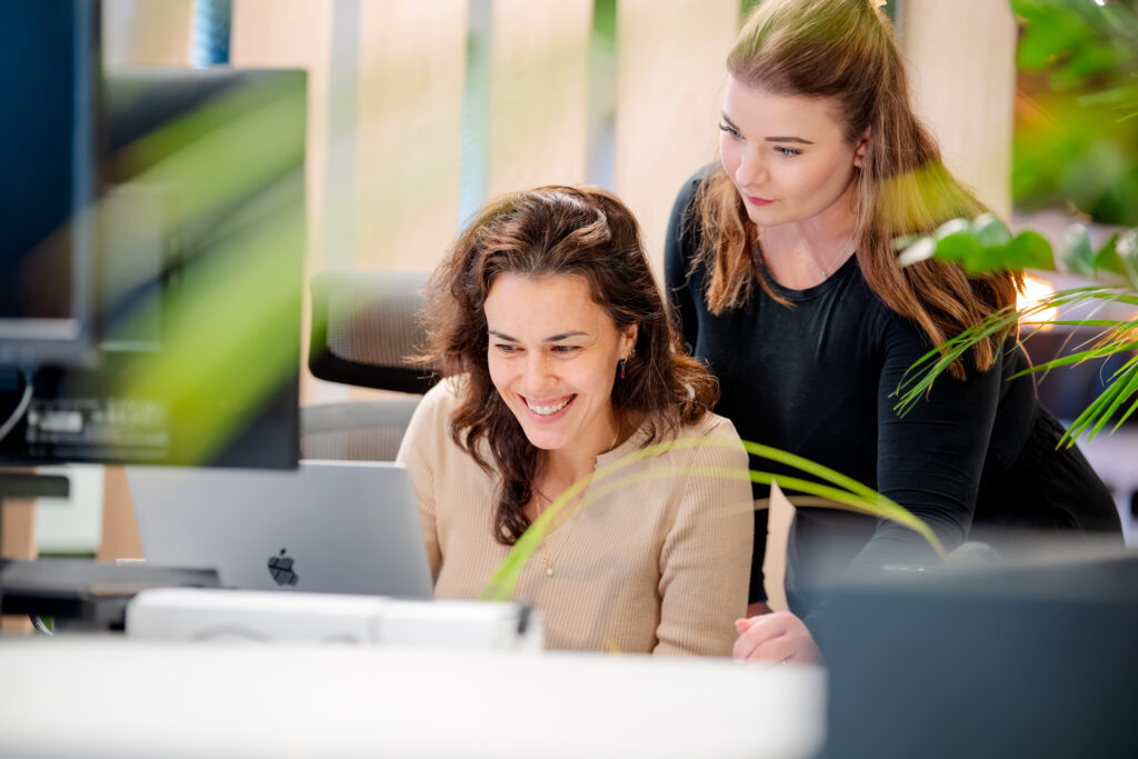 Twee vrouwen zitten achter een laptop in een kantooromgeving. Eén vrouw kijkt naar het scherm terwijl de ander naast haar sta