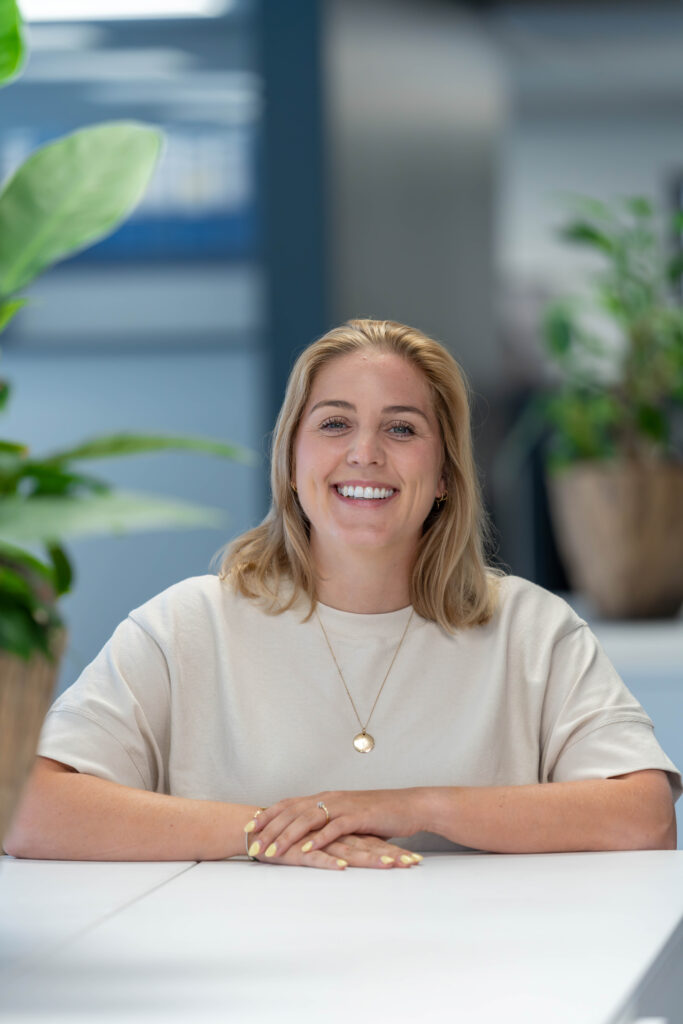 Een vrouw met kort, lichtbruin haar zit aan een witte tafel. Ze draagt een lichtbeige shirt en heeft haar handen op elkaar ge
