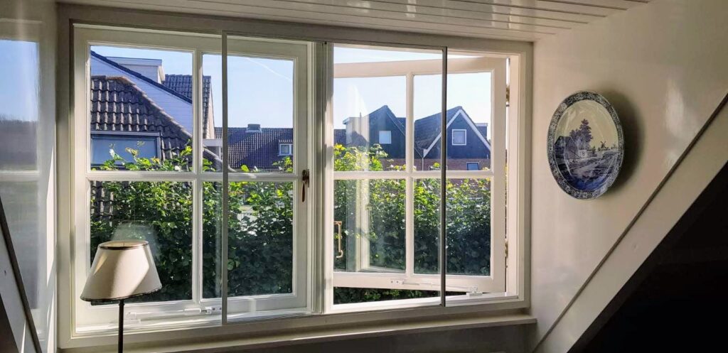 De afbeelding toont een open raam met een wit kozijn, waardoor een uitzicht op daken en struiken zichtbaar is. Aan de wand ha