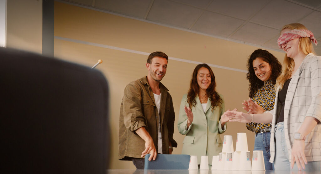 Vijf mensen staan samen rond een tafel in een ruimte. Eén persoon is geblinddoekt en probeert op een stapel plastic bekers te balanceren. De anderen kijken toe en lachen.